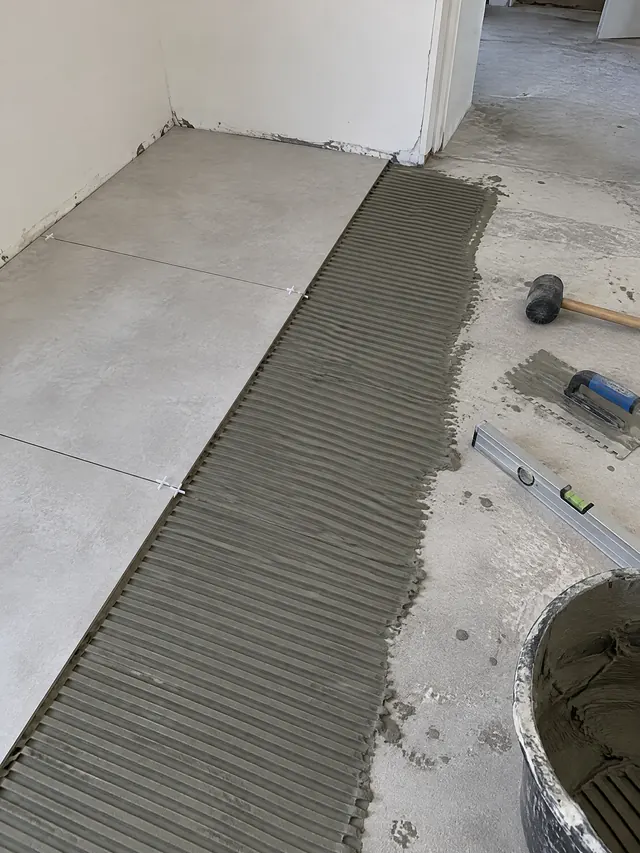Pose de carrelage au sol : colle peignée au peigne cranté sur la chape, grands carreaux gris posés avec croisillons, maillet et niveau au sol