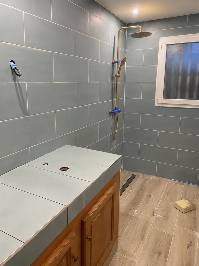 Douche à l’italienne en cours : faïence bleu gris posée, sol carrelé imitation bois en pente vers un caniveau inox, sorties de robinetterie protégées, paroi pas encore posée