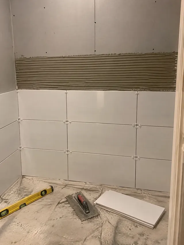 Mur de salle de bain en cours de faïence : carreaux blancs rectangulaires posés au croisillon sur la moitié basse, colle peignée en sillons droits au-dessus, niveau et peigne à colle au sol
