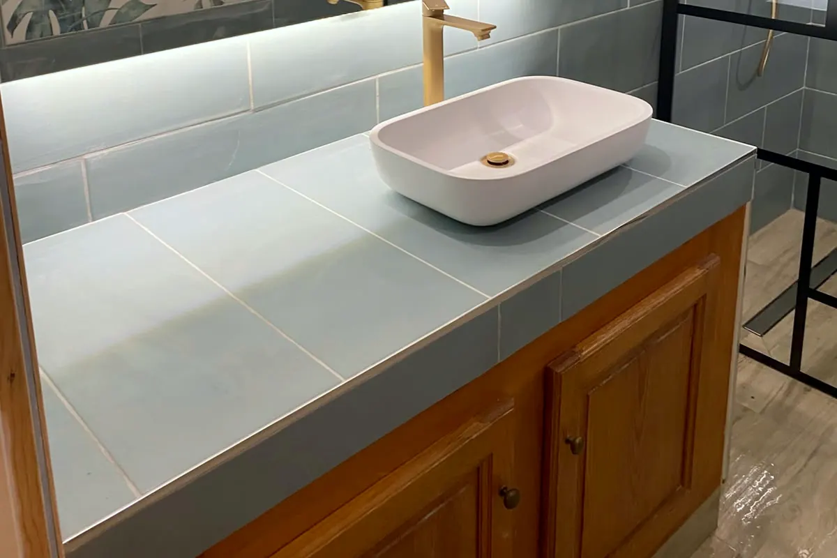 Salle de bain terminée : faïence bleu gris, plan de vasque carrelé, vasque blanche, panneau décoratif à motif de feuilles, paroi de douche noire