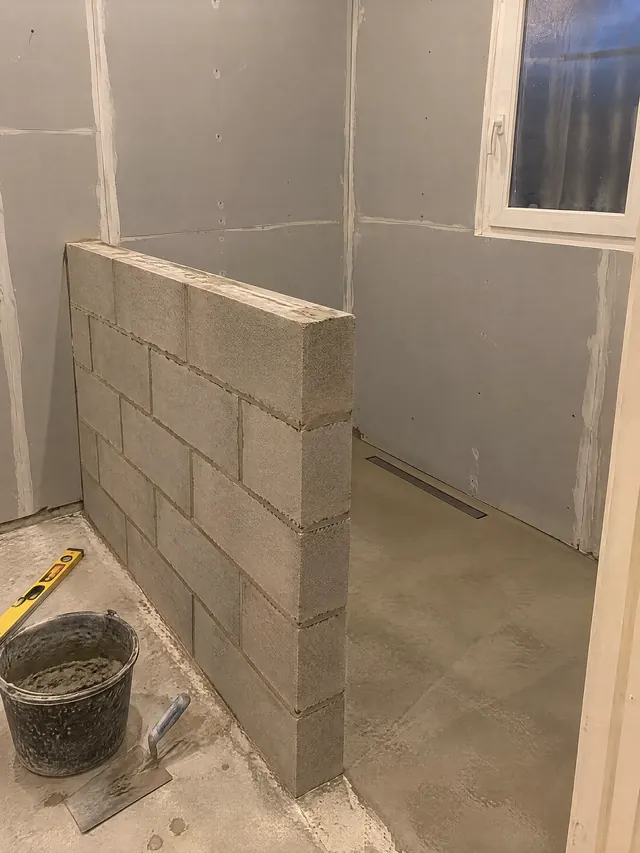 Salle de bain en travaux : muret en blocs béton monté au mortier pour fermer une future douche, sol de douche en chape avec caniveau, murs en plaques hydrofuges, seau de mortier et truelle