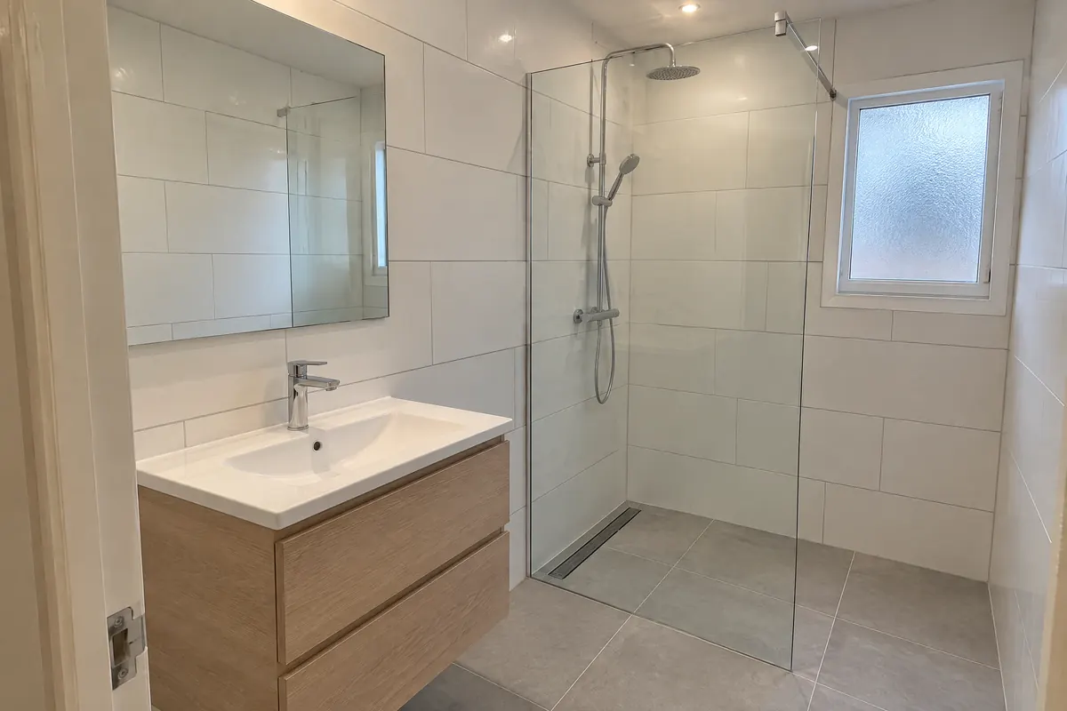 Petite salle de bain rénovée : faïence blanche rectangulaire, douche à l’italienne avec paroi vitrée et caniveau, carrelage gris clair, meuble vasque en chêne clair