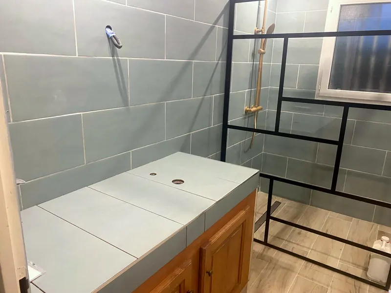 Salle de bain en cours : faïence bleu gris posée, plan de vasque carrelé percé pour le robinet, paroi de douche noire en place