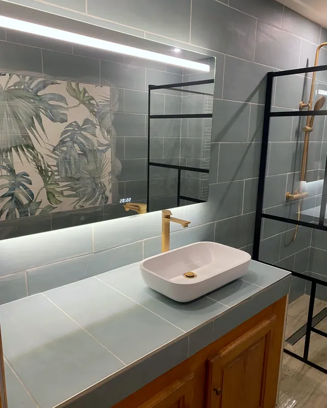 La même salle de bain terminée : vasque blanche, robinet laiton, miroir lumineux, panneau de carreaux à motif de feuilles